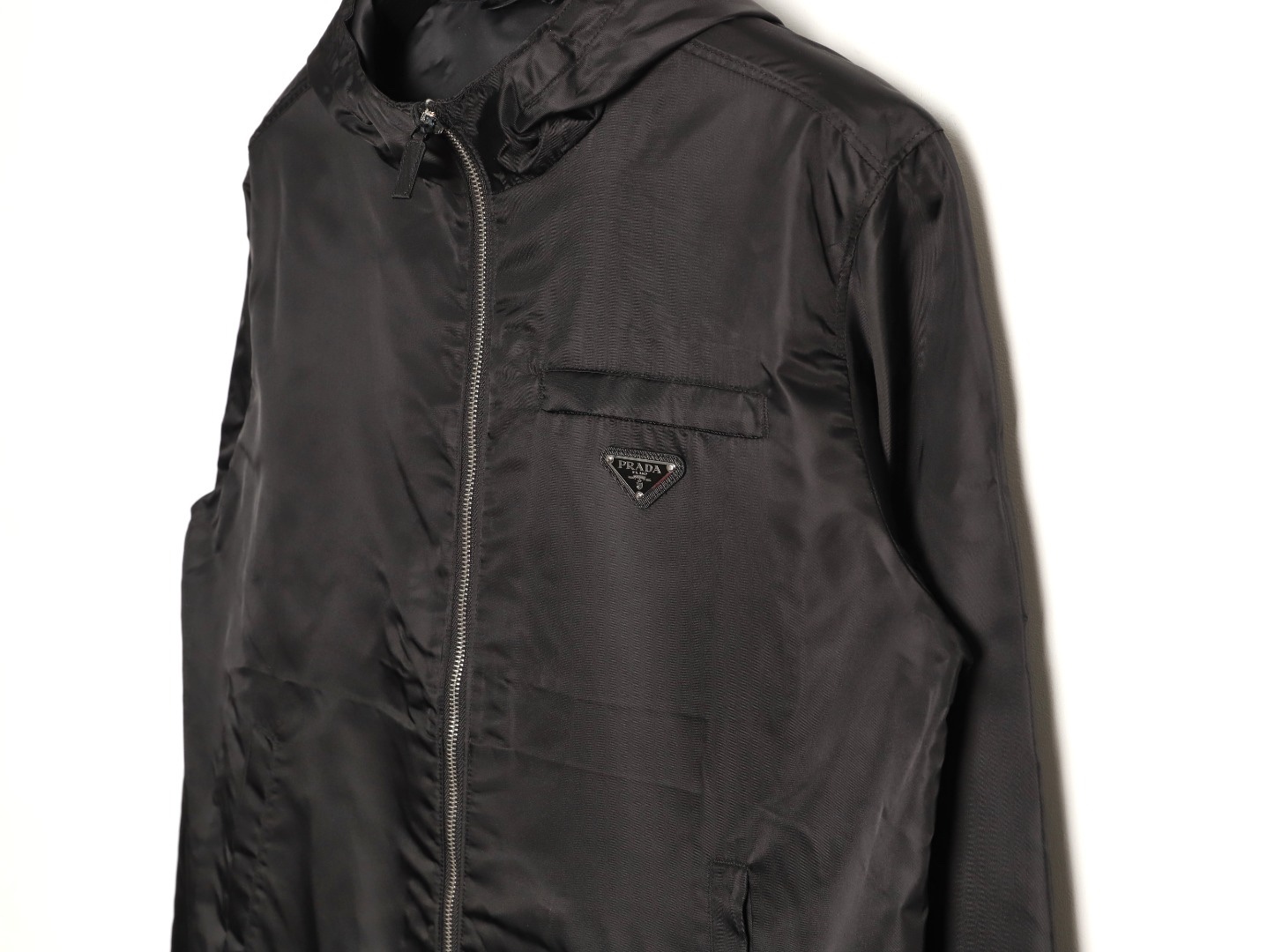🔝PRADA/普拉達PRD 拉鍊口袋尼龍連帽外套
選用品牌標誌性的再生尼龍面料，與傳統尼龍性能相當，具備高強度，抗撕裂，耐磨損等特性。其質地輕盈，同時又具備防水功能適合多場景需求。抗皺，快乾的特點也易於日常打理維護。
定制三角徽標通過4個0.8mm金屬爪釘固定於衣身，爪釘背面經手工打磨至圓潤，避免刮蹭。下擺彈力抽繩選用7芯高彈尼龍繩，直徑3mm，配合橡膠扣，可實現±5cm長度調節。抽繩孔採用「金屬雞眼+防磨環」雙重保護，雞眼內徑5mm，邊緣光滑無毛刺，反復抽拉500次無變形。
袖口採用紐扣調