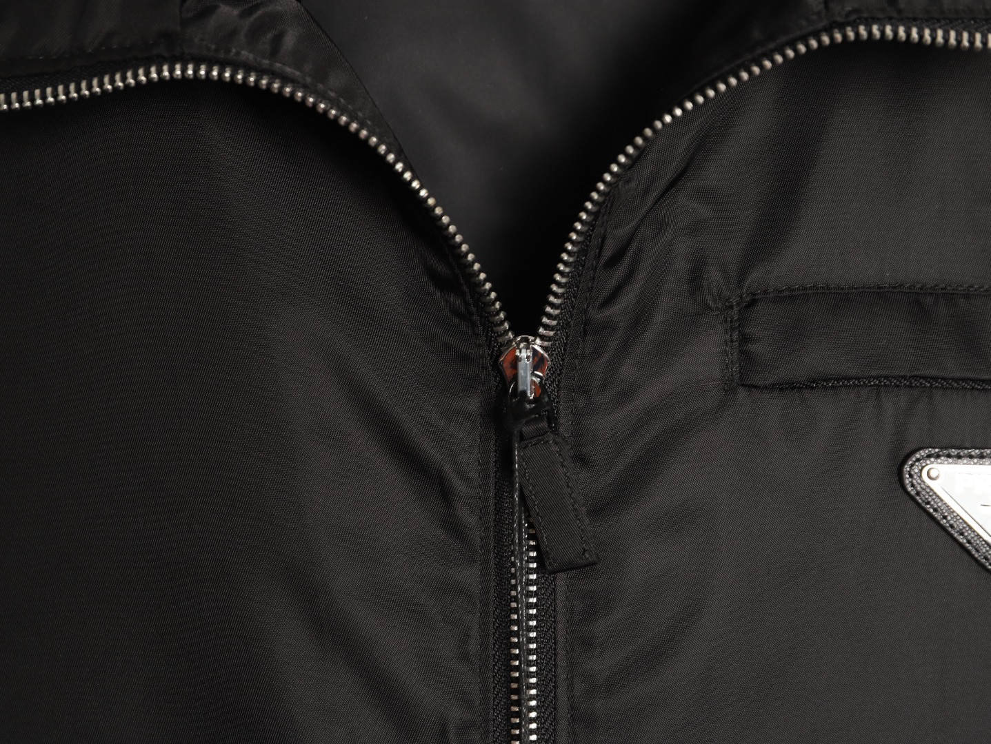 🔝PRADA/普拉達PRD 拉鍊口袋尼龍連帽外套
選用品牌標誌性的再生尼龍面料，與傳統尼龍性能相當，具備高強度，抗撕裂，耐磨損等特性。其質地輕盈，同時又具備防水功能適合多場景需求。抗皺，快乾的特點也易於日常打理維護。
定制三角徽標通過4個0.8mm金屬爪釘固定於衣身，爪釘背面經手工打磨至圓潤，避免刮蹭。下擺彈力抽繩選用7芯高彈尼龍繩，直徑3mm，配合橡膠扣，可實現±5cm長度調節。抽繩孔採用「金屬雞眼+防磨環」雙重保護，雞眼內徑5mm，邊緣光滑無毛刺，反復抽拉500次無變形。
袖口採用紐扣調